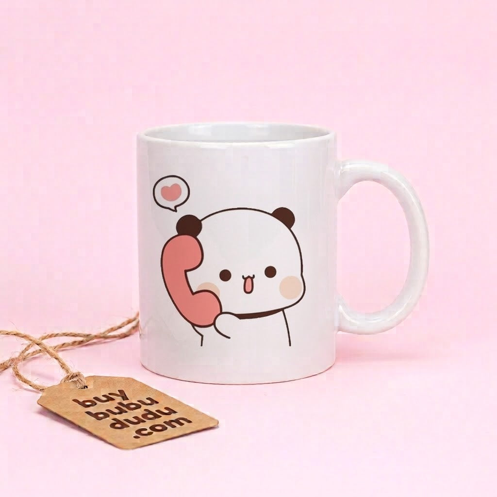 A white ceramic mug with a cute cartoon panda holding a pink phone, set against a solid pink background. A brown paper tag attached to twine is in the foreground.