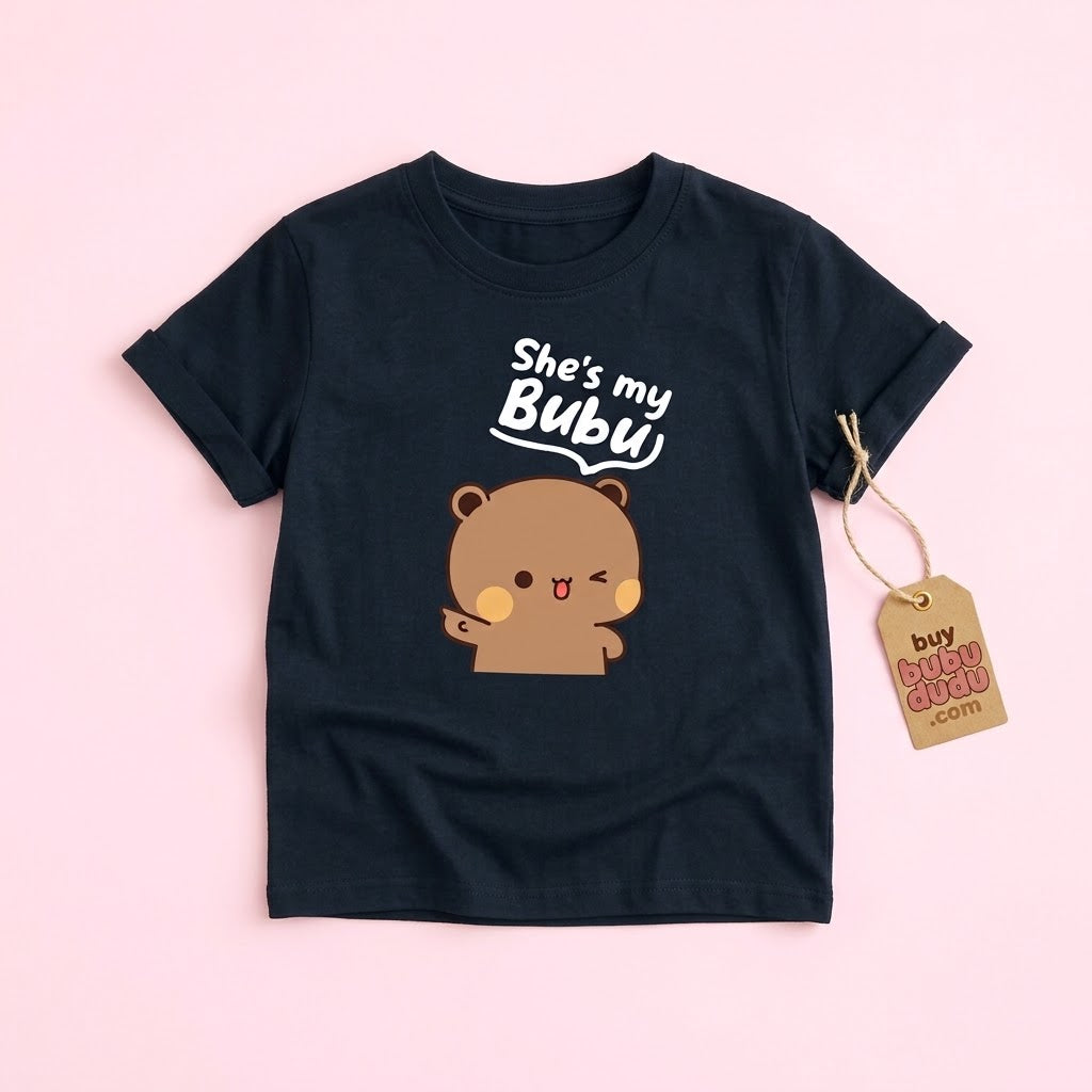A black short-sleeve t-shirt with a cute cartoon bear graphic and the text 'She's my Bublu' on the front, displayed flat on a light pink background with a paper hangtag attached.