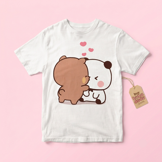 A white short-sleeve t-shirt with a cute cartoon graphic of a brown bear hugging a white panda under pink hearts, displayed on a solid pink background with a paper hangtag.