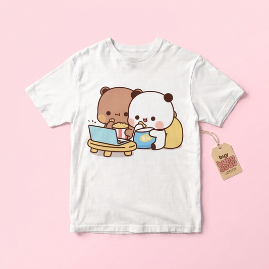 A white short-sleeve t-shirt with a cute cartoon graphic of a brown bear eating popcorn and a white panda drinking from a blue cup. The shirt is displayed flat on a light pink background with a paper hangtag attached to the right sleeve.