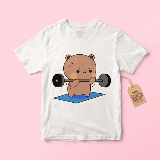 A white short-sleeve t-shirt with a cartoon graphic of a brown bear lifting a barbell on a blue mat, displayed on a light pink background with a paper hangtag attached.